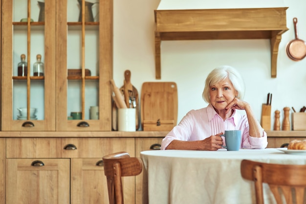 Donna anziana allegra con una tazza di bevanda calda seduta in cucina