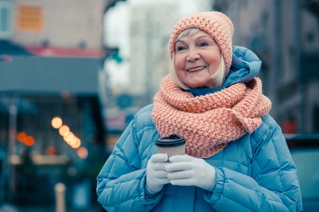 Donna anziana allegra in abiti invernali in piedi all'aperto con una tazza di caffè in cartone tra le mani e sorridente felicemente