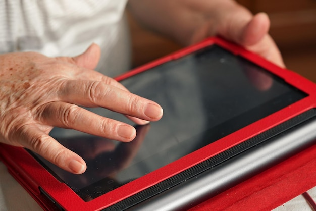 Donna anziana anziana che lavora con il tablet del computer in custodia protettiva rossa, dettaglio del primo piano sullo schermo commovente della mano