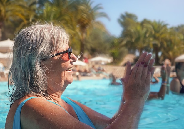 Donna anziana anziana con i capelli grigi, che indossa un costume da bagno blu che fa aerobica in acqua nella piscina dell'hotel, battendo le mani