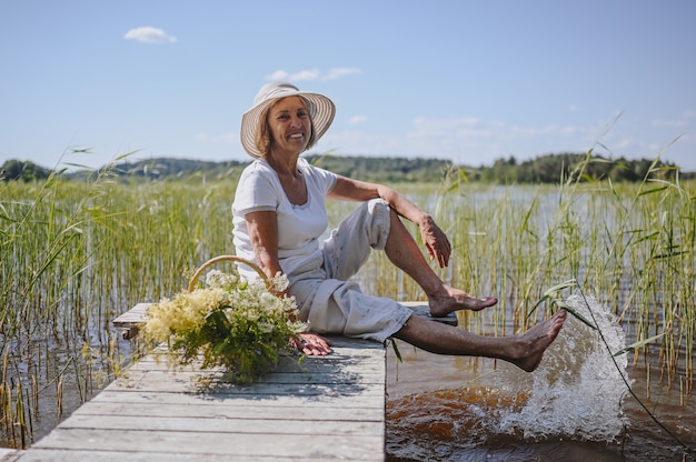 Donna anziana anziana sorridente felice in cappello di paglia che si siede sul pilastro di legno dal lago con i fiori in