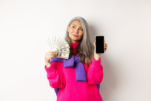 Donna anziana asiatica alla moda che mostra dollari di denaro e schermo vuoto dello smartphone, dimostra il negozio online, in piedi su sfondo bianco