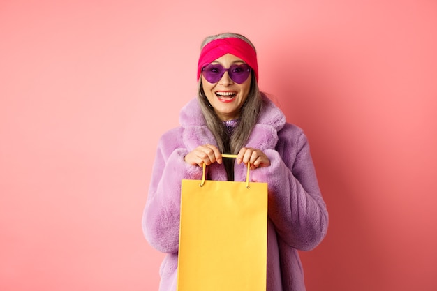 Donna anziana asiatica alla moda che va a fare shopping con occhiali da sole e vestiti invernali alla moda, tiene in mano un sacchetto di carta dal negozio e sorride felice alla telecamera, sfondo rosa.