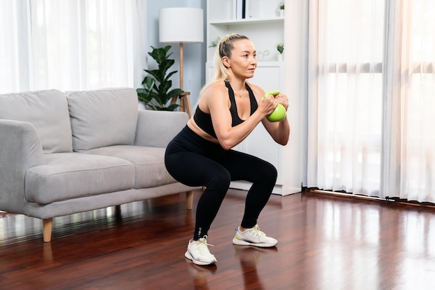 Donna anziana atletica e sportiva impegnata in una sessione di allenamento per le gambe con esercizi di squat e palla con bollitore a corpo libero a casa come concetto di stile di vita sano e in forma dopo il pensionamento