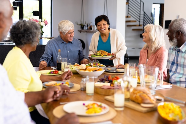 Donna anziana biraziale sorridente che serve cibo ad amici multirazziali al tavolo da pranzo in una casa di cura. Pasto, cibo, bevande, felice, inalterato, insieme, sostegno, vita assistita e concetto di pensionamento.