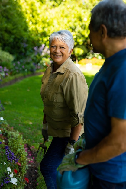 Donna anziana birazziale sorridente che guarda il marito che tiene l'annaffiatoio mentre si trova in cortile
