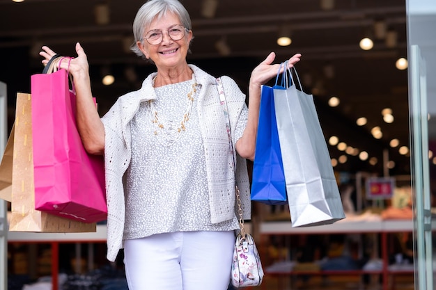 Donna anziana caucasica attraente che tiene le borse della spesa mentre esce da un negozio godendo del concetto di consumismo dello shopping