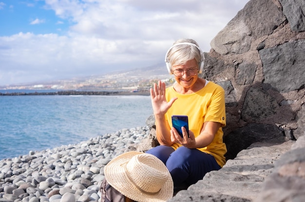 Donna anziana caucasica che indossa le cuffie seduta su una spiaggia di ciottoli in mare utilizzando il telefono cellulare Donna matura che tiene il cellulare utilizzando tecnologia e sociale Concetto di libertà di vacanza