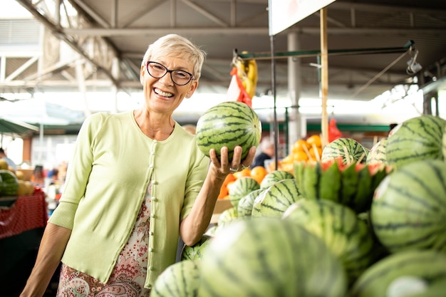 donna anziana che acquista angurie fresche e frutta al mercato e tiene in mano una borsa piena di cibo sano