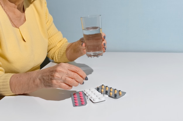 Donna anziana che assume farmaci. Chiuda in su delle mani femminili che tengono bicchiere d'acqua e pillole. Concetto di trattamento, pensionamento per malattia