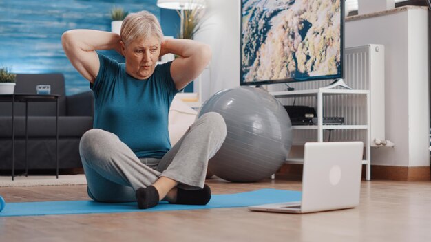 Donna anziana che guarda un video di formazione online sul laptop e fa esercizi di ginnastica mentre è seduta su un tappetino da yoga. Adulto in pensione che guarda la lezione di allenamento sul computer per l'attività di fitness.