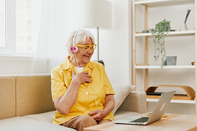 Donna anziana che indossa le cuffie con un computer portatile comunicazione videochiamata online sorriso felicità seduto sul divano a casa e lavorando in una camicia gialla lo stile di vita di una donna in pensione