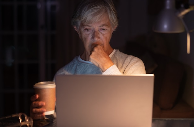 Donna anziana che sfoglia i contenuti dei social media utilizzando il computer portatile sulla scrivania con in mano una tazza di caffè. Vecchia donna in pensione di notte che trascorre il suo tempo lavorando o studiando sul laptop