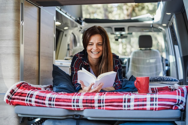 Donna anziana che si diverte all'interno del camper mentre legge un libro e beve caffè - Focus on face