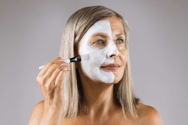 Donna anziana che utilizza un pennello cosmetico per applicare la maschera di argilla bianca