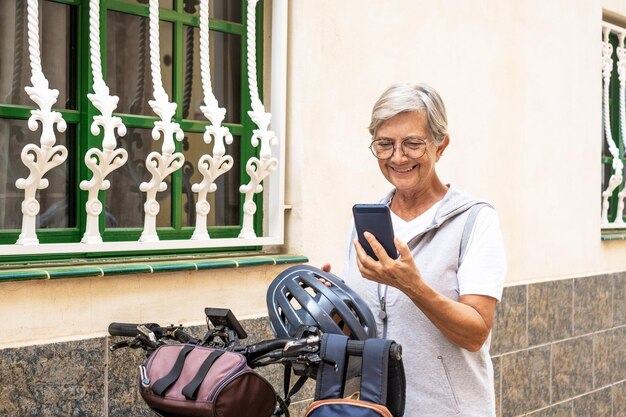 Donna anziana ciclista allegra in strada urbana che tiene il casco prima di correre con la sua bicicletta elettrica utilizzando il telefono cellulare Concetto di stile di vita sano e mobilità sostenibile