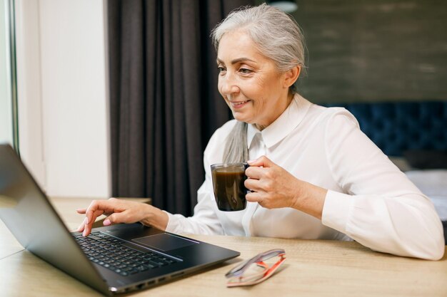 Donna anziana con capelli bianchi che beve caffè mentre si lavora con il computer portatile