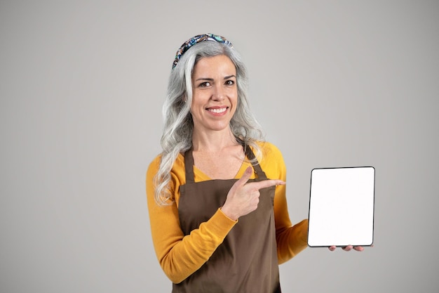 Donna anziana con capelli grigi in grembiule che tiene una tavoletta che offre consigli sui prodotti agricoli