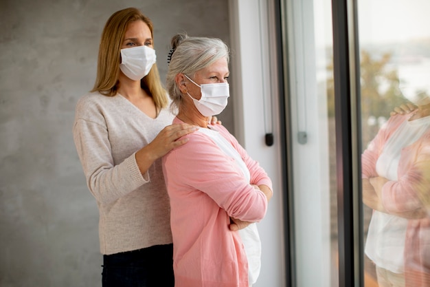 Donna anziana con figlia premurosa a casa che indossa maschere mediche come protezione dal coronavirus