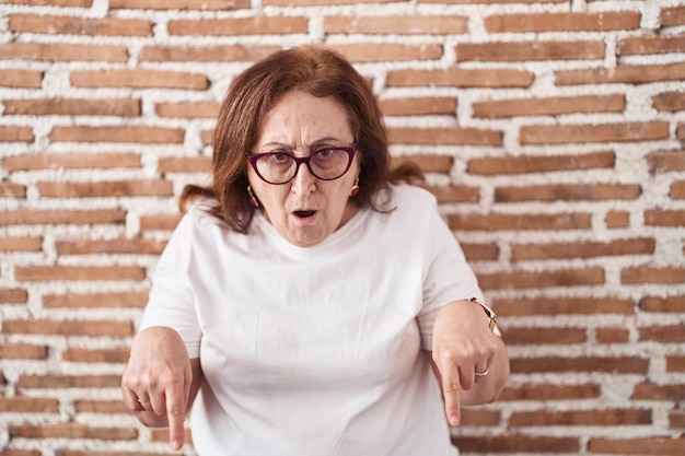 Donna anziana con gli occhiali in piedi sopra il muro di mattoni rivolto verso il basso con le dita che mostrano pubblicità, faccia sorpresa e bocca aperta