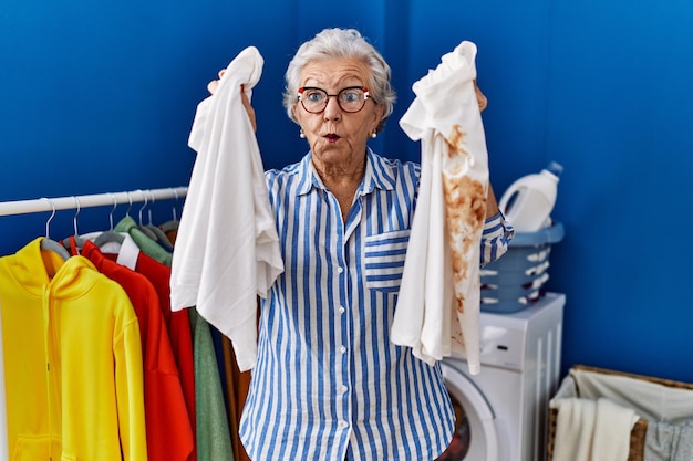 Donna anziana con i capelli grigi che tiene una maglietta bianca pulita e una maglietta con una macchia sporca che fa la faccia da pesce con la bocca e gli occhi strabici, pazza e comica.