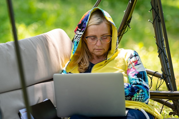 Donna anziana con laptop e documenti che lavorano in giardino sul divano a dondolo concetto di home office verde