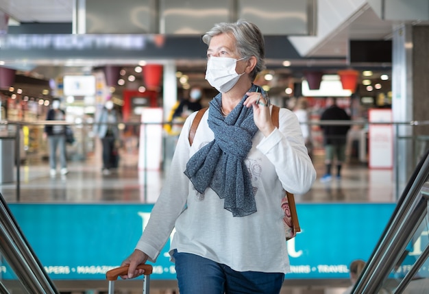Donna anziana con maschera facciale nell'area duty-free dell'aeroporto con bagagli in attesa della partenza del volo