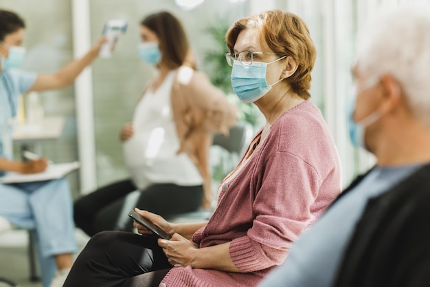 Donna anziana con maschera facciale seduta in una sala d'attesa di un ospedale prima della vaccinazione Covid-19.