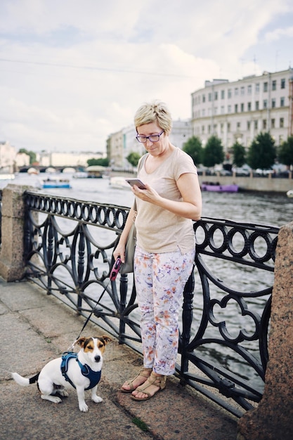 Donna anziana con un cane al guinzaglio e guardando il suo telefono vicino al fiume in una strada cittadina