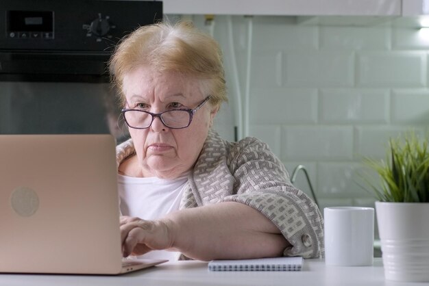 Donna anziana concentrata seduta al tavolo a casa che lavora utilizzando computer portatile con faccia seria