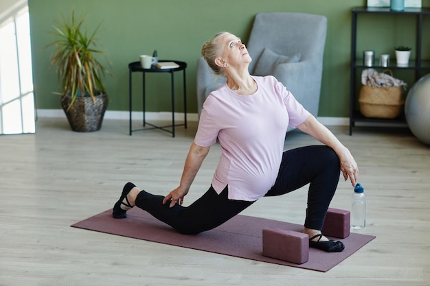 Donna anziana contemporanea in maglietta e leggins che guarda verso l'alto durante la pratica dell'esercizio yoga sul tappetino in soggiorno