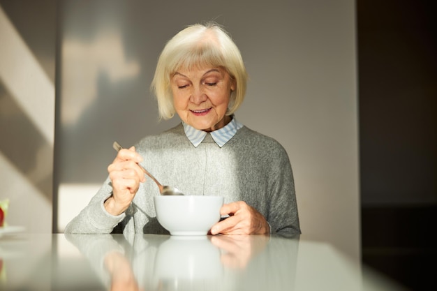 Donna anziana contenta che fa colazione nella cucina illuminata dal sole