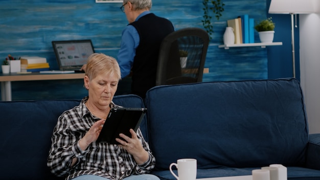 Donna anziana di mezza età che si rilassa mentre legge tablet e libro seduto sul divano di casa, mentre un uomo adulto anziano lavora al computer portatile in background. Persona che si diverte a utilizzare il blocco note mentre naviga in Internet per lo shopping