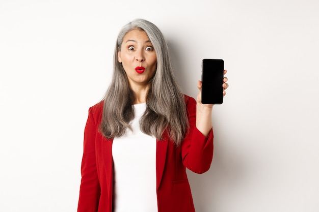 Donna anziana divertita in blazer rosso, che sembra affascinata e mostra lo schermo vuoto dello smartphone, in piedi su sfondo bianco.