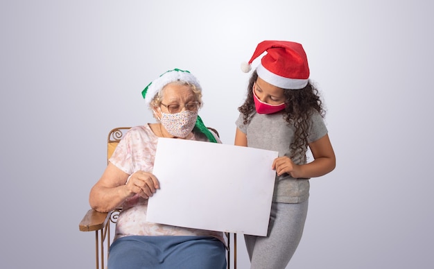 Donna anziana e bambino con cappello di Natale e maschera con cartello bianco su grigio