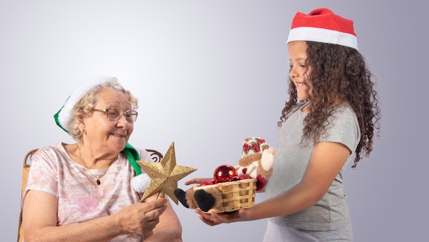 Donna anziana e bambino con cappello di Natale e ornamenti natalizi nelle mani su grigio