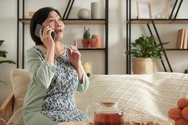 Donna anziana emotiva eccitata che parla al telefono con un amico quando è seduta su un comodo divano a casa