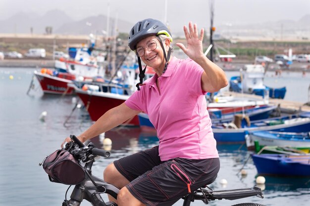 Donna anziana felice attiva con casco che corre sulla sua bicicletta elettrica lungo il porto marittimo guardando la fotocamera Concetto di libertà e vacanze