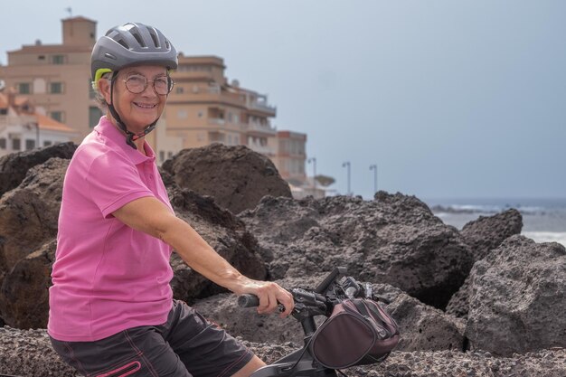 Donna anziana felice attiva con casco che esegue la sua bicicletta elettrica lungo il mare guardando la fotocamera