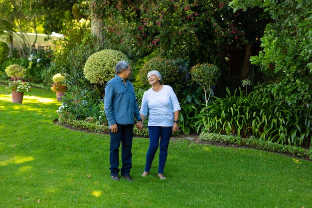 Donna anziana felice birazziale che tiene la mano del marito mentre si trova contro piante e alberi nel parco