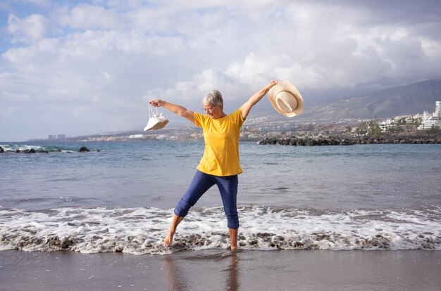 Donna anziana felice con le braccia tese sulla riva del mare che tiene scarpe e cappello in mano Nonna anziana a piedi nudi nell'acqua godendosi la vacanza e la libertà