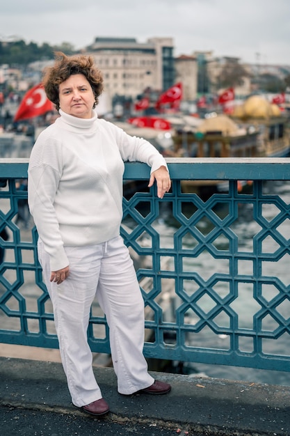 Donna anziana in abiti bianchi sul ponte di Galata in una giornata nuvolosa. bandiere turche sventolano sullo sfondo. una donna di 50 anni, bassa, guarda nella telecamera, all'aperto, foto verticale