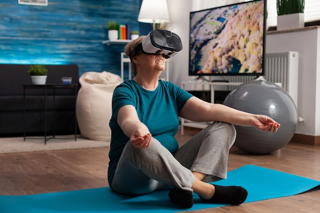 Donna anziana in buona salute che usa le cuffie per realtà virtuale meditando mentre è seduta nella posizione del loto sul tappetino da yoga in soggiorno durante l'allenamento di meditazione meditation