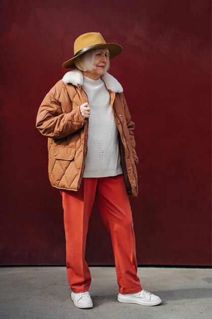 Donna anziana in capispalla alla moda e cappello che guarda lontano mentre si trova contro il muro vinoso.