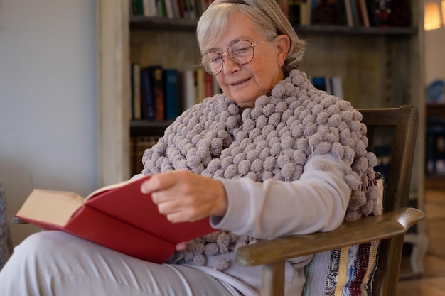 Donna anziana in occhiali seduto sulla poltrona che legge il romanzo bestseller del libro cartaceo Pensionamento per il tempo libero