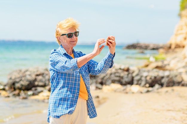 Donna anziana in piedi sulla spiaggia e scattare foto su un telefono cellulare