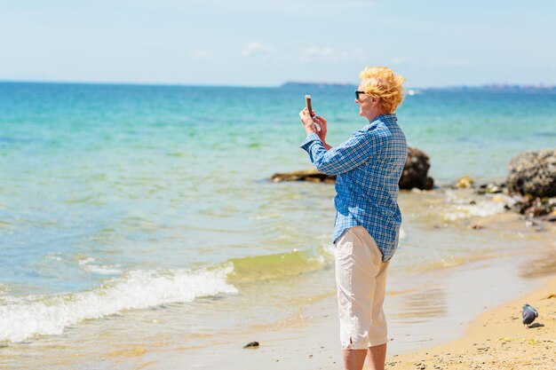 Donna anziana in piedi sulla spiaggia e scattare foto su un telefono cellulare