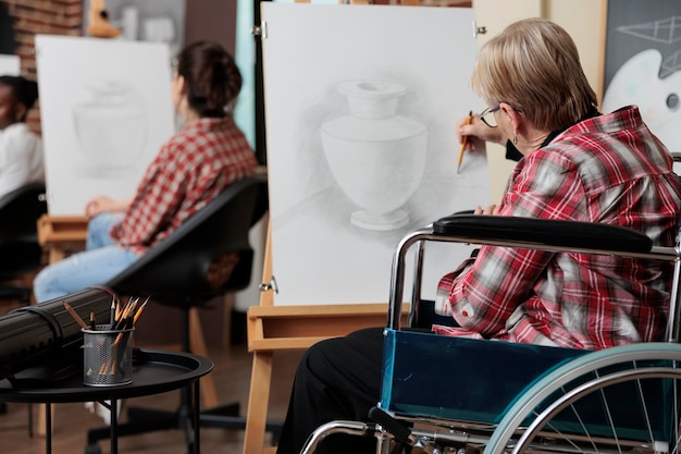 Donna anziana in sedia a rotelle che presta attenzione alla classe d'arte per la riabilitazione che disegna uno schizzo di vaso su tela bianca sviluppando abilità di schizzo per la crescita personale. Gruppo eterogeneo di persone che creano opere d'arte moderne