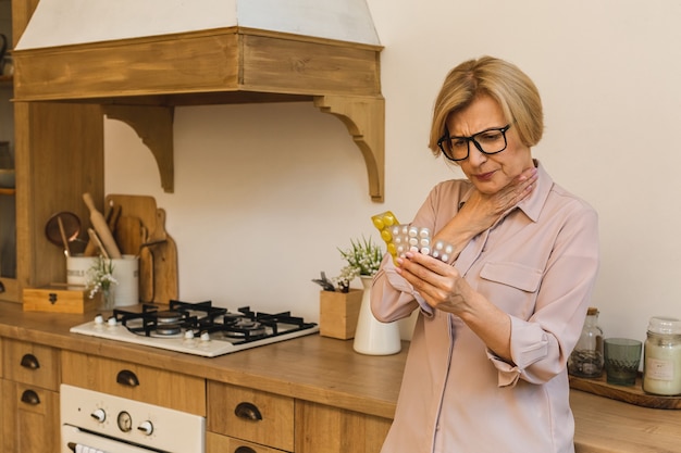 Donna anziana invecchiata matura che tiene le vesciche con le pillole nella cucina a casa. Sentirsi male.