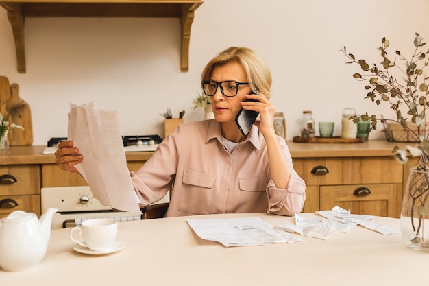 Donna anziana matura di mezza età che tiene una fattura cartacea o una lettera a casa per effettuare pagamenti online sul sito Web sul telefono cellulare, calcolare il costo delle tasse finanziarie, rivedere il conto bancario.